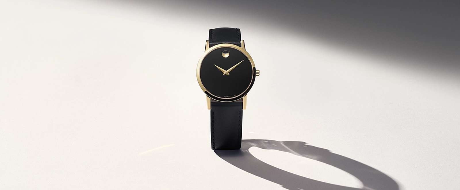 Reloj Movado Museum Classic dorado con correa de cuero negro y su sombra sobre fondo claro
