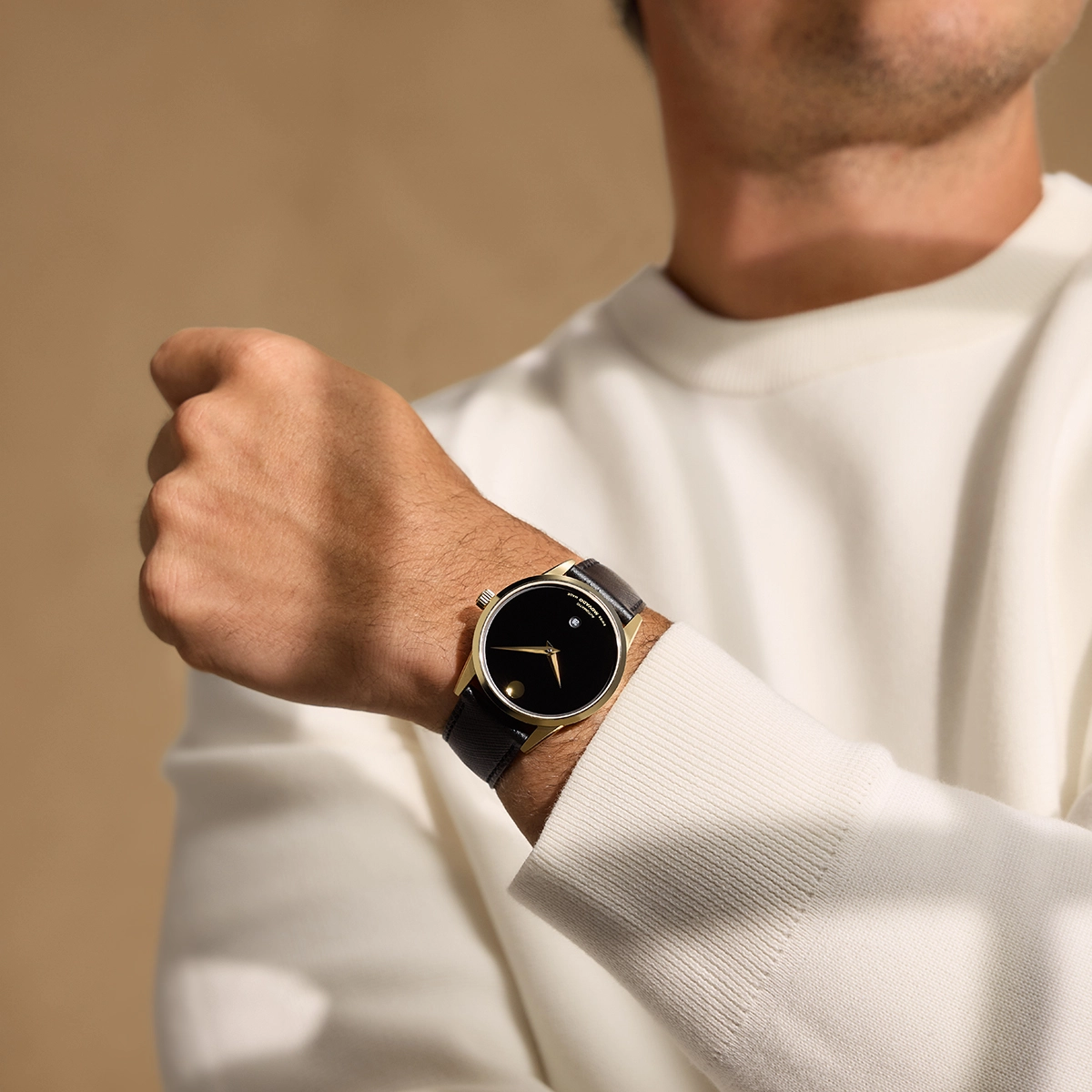 Hombre de suéter blanco luciendo un Movado Museum Classic dorado con correa negra